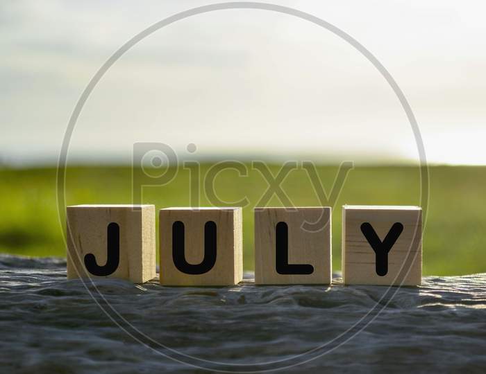 July Text On Wooden Cube Block On Old Tree Stump With Blurred Background Of Green Grass Ocean And Sunset