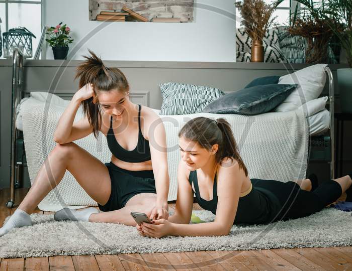 Young Women Go In For Sports At Home, Workout Online. Two Athletes Are Stretching, Meditating, Discussing Workout In The Bedroom, In The Background There Is A Bed, A Vase, A Carpet.