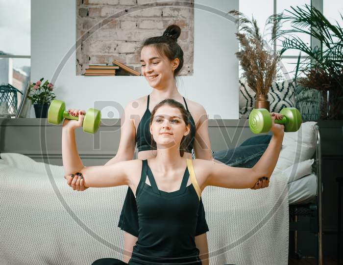A Strong And Beautiful Sports Fitness Girls In Sportswear  Doing Hand Biceps Exercise With Dumbbells In Their Bright And Airy Bedroom With A Minimalistic Interior.