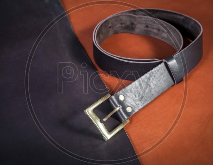 A Close-Up Black Belt Made Of Genuine Leather Made By Hand And A Few Buckles Lie On The Table On Pieces Of Black And Brown Leather