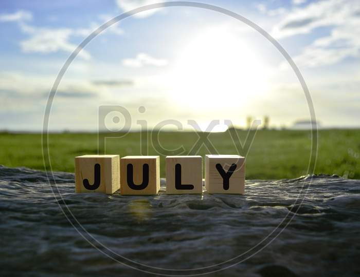 July Text On Wooden Cube Block On Old Tree Stump With Blurred Background Of Green Grass Ocean And Sunset