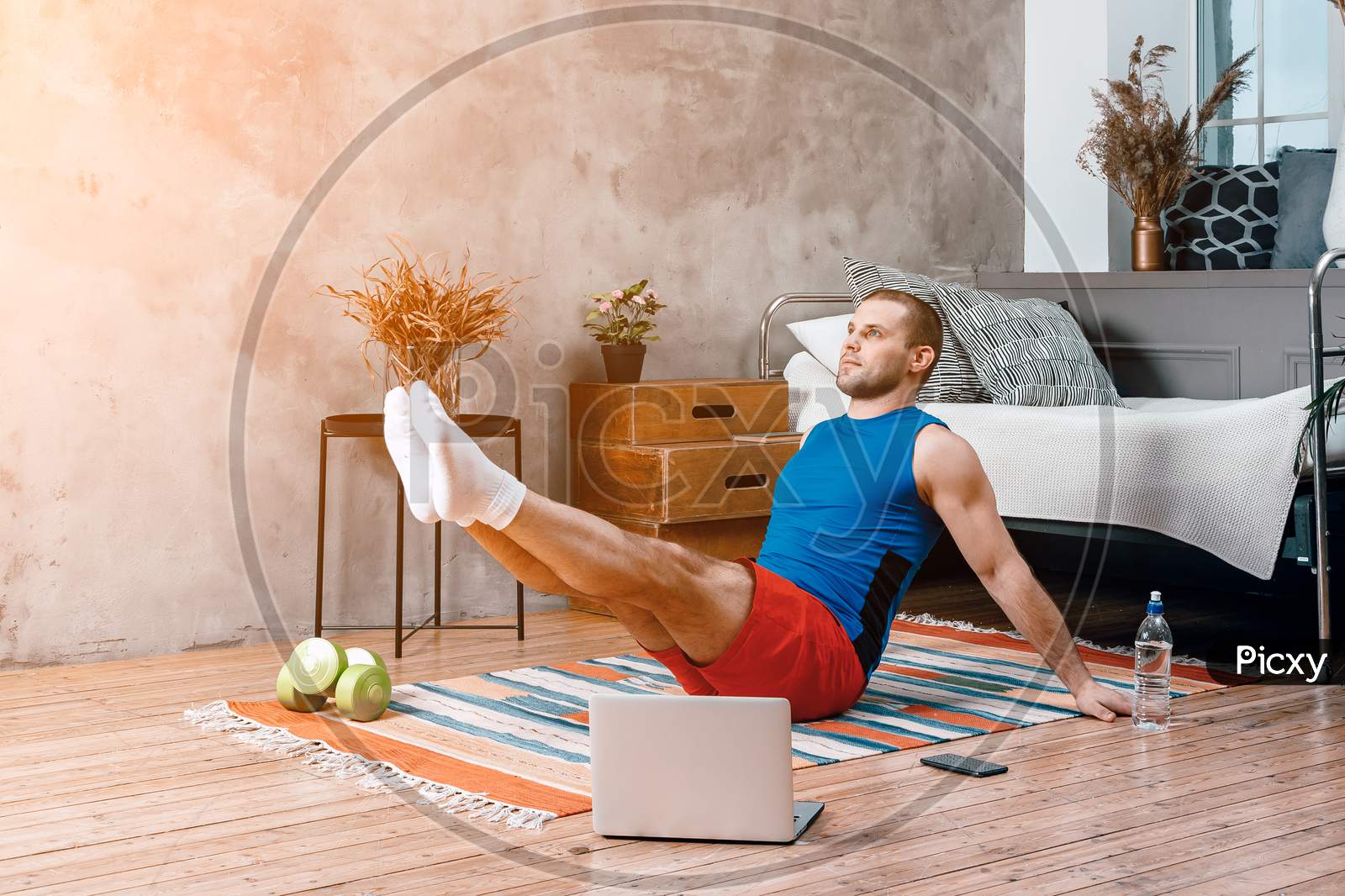 Young Man Goes In For Sports At Home, Training Online. The Athlete Makes The Press, Lifting His Legs Up,  Watches A Movie And  Social Networks  On Laptop