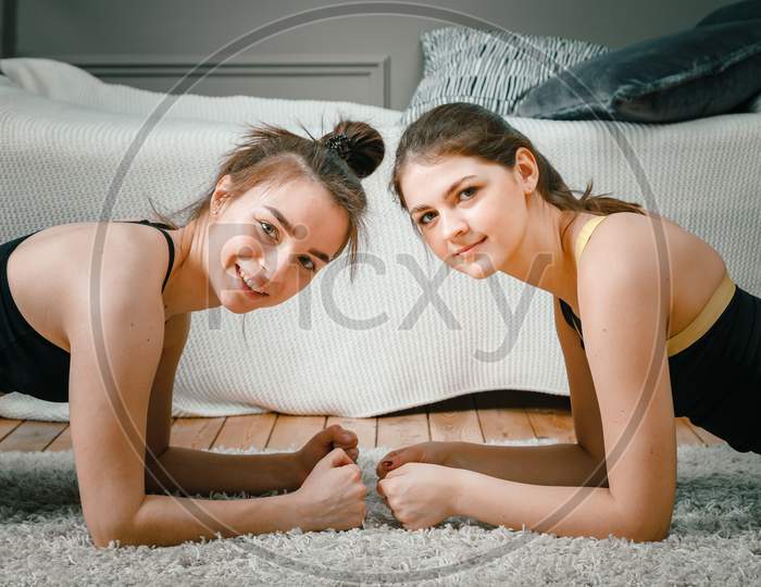 The Young Women Goes In For Sports At Home. Portrait Of The Two  Cheerful Sporty Women  Makes  Plank In The Bedroom