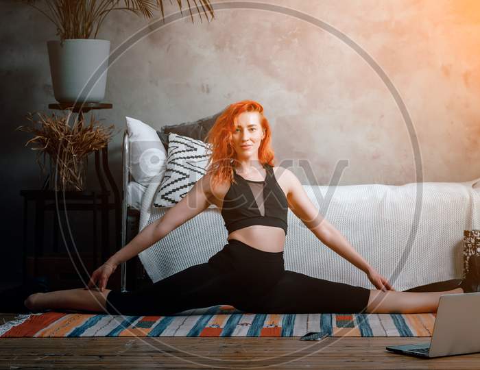 A Young Woman Goes In For Sports At Home, Online Workout . The Athlete  Stretching , Meditating, Sitting On A Twine  In The Bedroom, In The Background There Is A Bed, A Vase, A Carpet.