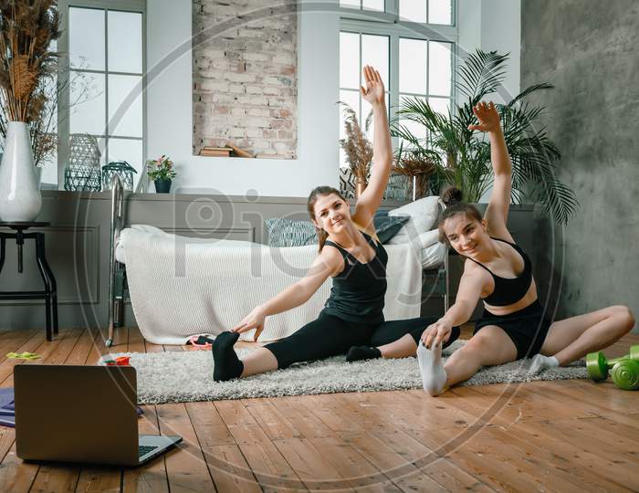 Young Women Go In For Sports At Home, Workout Online. Two Athletes Are Stretching, Meditating  In The Bedroom, In The Background There Is A Bed, A Vase, A Carpet.