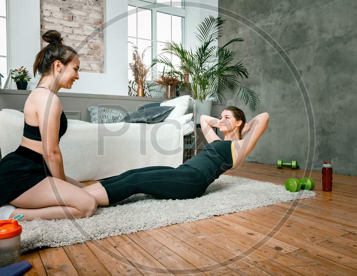 Two Women Friends At Home Chatting, Smiling And Makes Sports. Cheerful Athletic Woman Presses, And The Second Sits Nearby And Monitors Correct Execution In The Bedroom