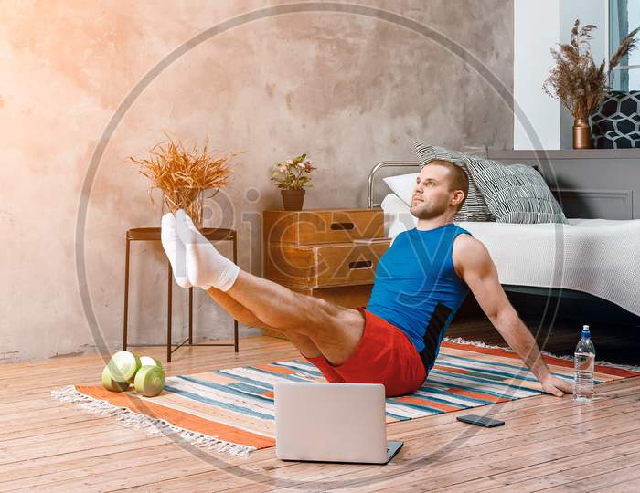 Young Man Goes In For Sports At Home, Training Online. The Athlete Makes The Press, Lifting His Legs Up,  Watches A Movie And  Social Networks  On Laptop