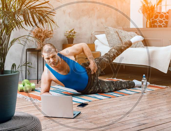 The Young Man Goes In For Sports At Home. Cheerful Sportsman With Black Hair Shakes Leg, Lunges And Watching Online Workout From Laptop In Bedroom