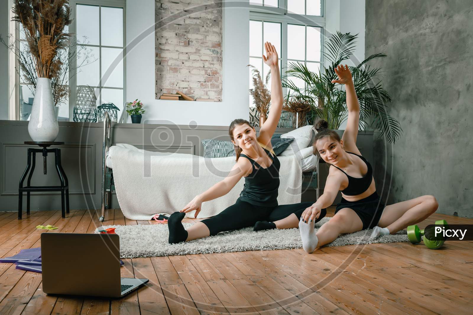 Young Women Go In For Sports At Home, Workout Online. Two Athletes Are Stretching, Meditating  In The Bedroom, In The Background There Is A Bed, A Vase, A Carpet.