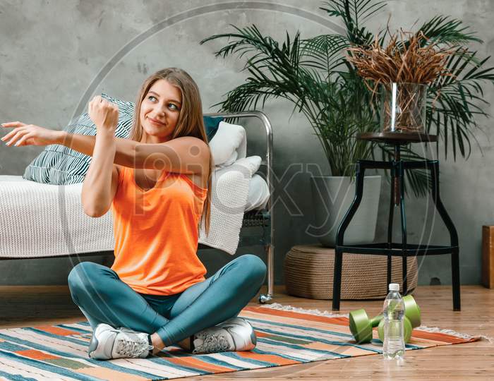 A Young Woman Goes In For Sports At Home, Online Workout . The Athlete  Stretching  Arm, Meditating, Sitting On A Floor  In The Bedroom