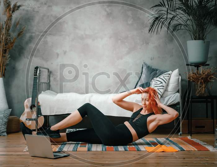 The Young Woman Goes In For Sports At Home. Cheerful Sport Woman With Red Hair Makes The Press In The Bedroom, Next To There Is A Laptop With Online Training