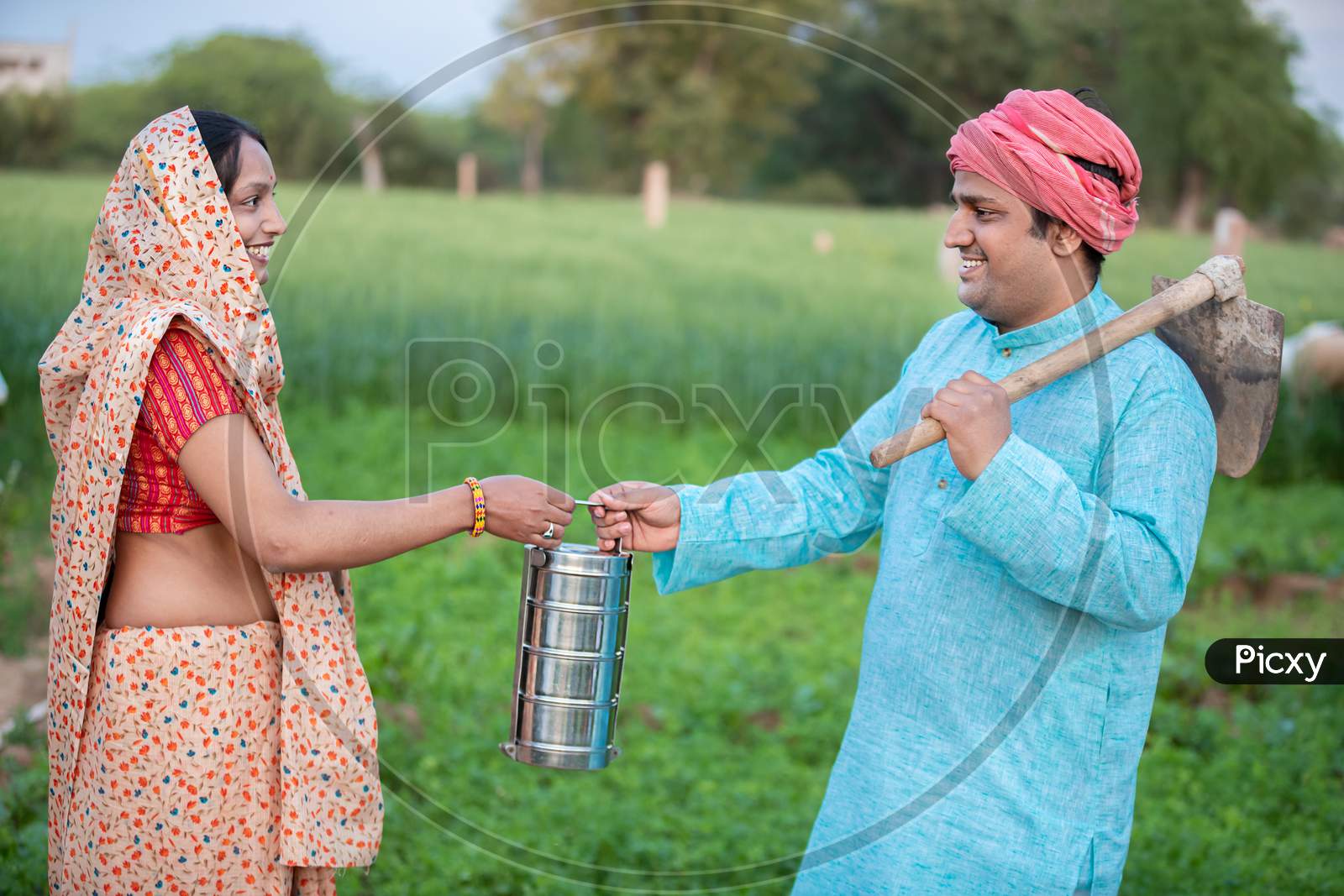 Happy Traditional Indian Woman Wearing Sari Giving Tiffin Box To His Husband In Agriculture Field, Happy Farmer Couple Holding Lunch Box.