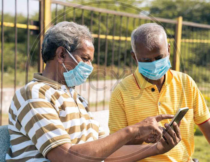 Side View Selective Focus On Mobile, Elderly Man In Medical Face Mask Showing Mobile Phone To His Old Friend At Park - Concept Of Senior People Using Smartphone, Technology, Internet And Social Media