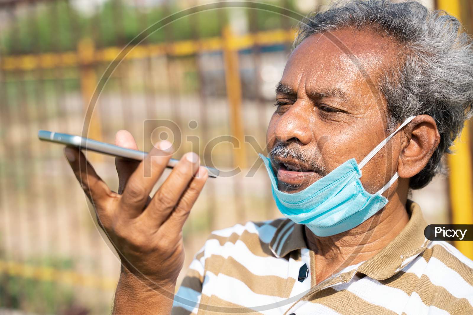 Senior Man With Medical Face Mask Below The Jaw Talking On Mobile Phone At Park - Concept Of Improper Mask Use Due To Coronavirus Covid-19 Pandemics.