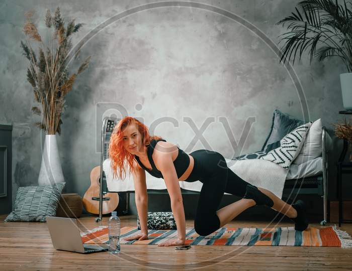 A Young Woman Goes In For Sports At Home, Online Workout From The Laptop. The Athlete Lunges, Watches A Movie  In The Bedroom, In The Background There Is A Bed, A Vase, A Carpet.