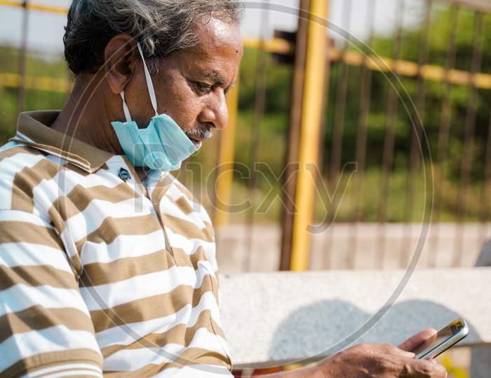Senior Man With Medical Face Mask Below The Jaw Using Smartphone At Park - Concept Of Improper Mask Use Due To Coronavirus Covid-19 Pandemics.