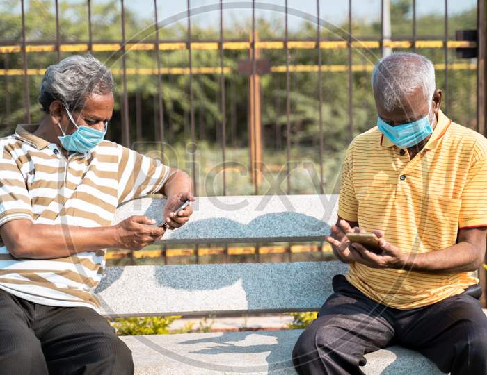 Two Elderly People With Medical Face Mask Busy Using Mobile Phone While Sitting At Park While Maintaining Social Distance - Concept Seniors Using Smartphone, Technology, Social Media And Apps