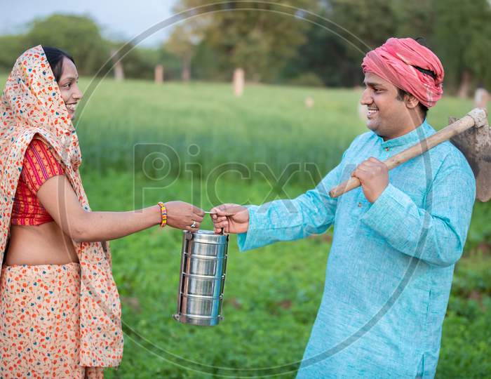 Happy Traditional Indian Woman Wearing Sari Giving Tiffin Box To His Husband In Agriculture Field, Happy Farmer Couple Holding Lunch Box.