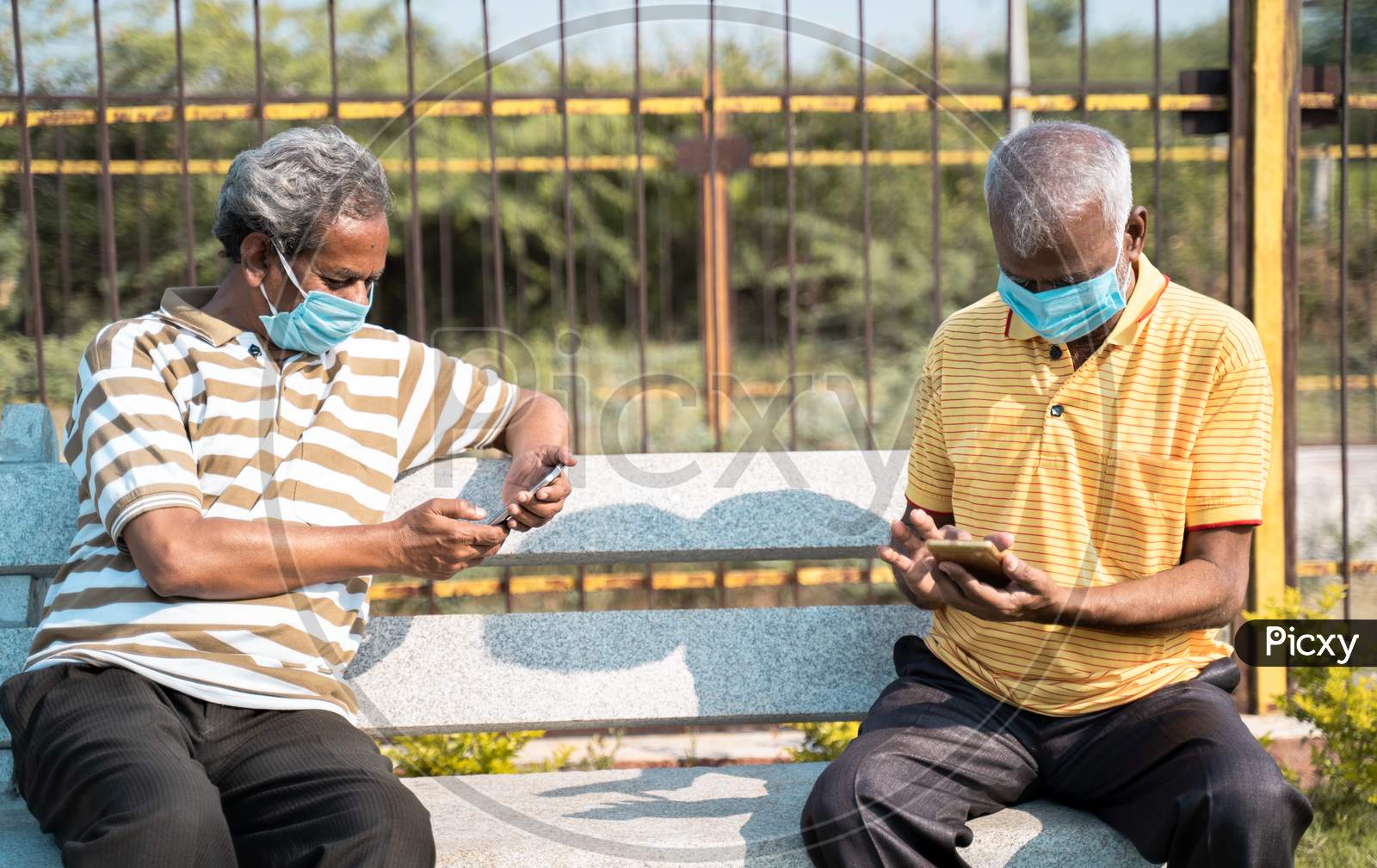 Two Elderly People With Medical Face Mask Busy Using Mobile Phone While Sitting At Park While Maintaining Social Distance - Concept Seniors Using Smartphone, Technology, Social Media And Apps