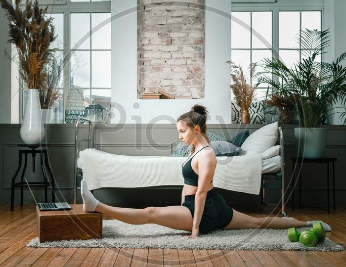 A Strong And Beautiful Sports Fitness Girl In Sportswear Stretching And Doing  Twine  In Her Bright And Airy Bedroom With A Minimalistic Interior.