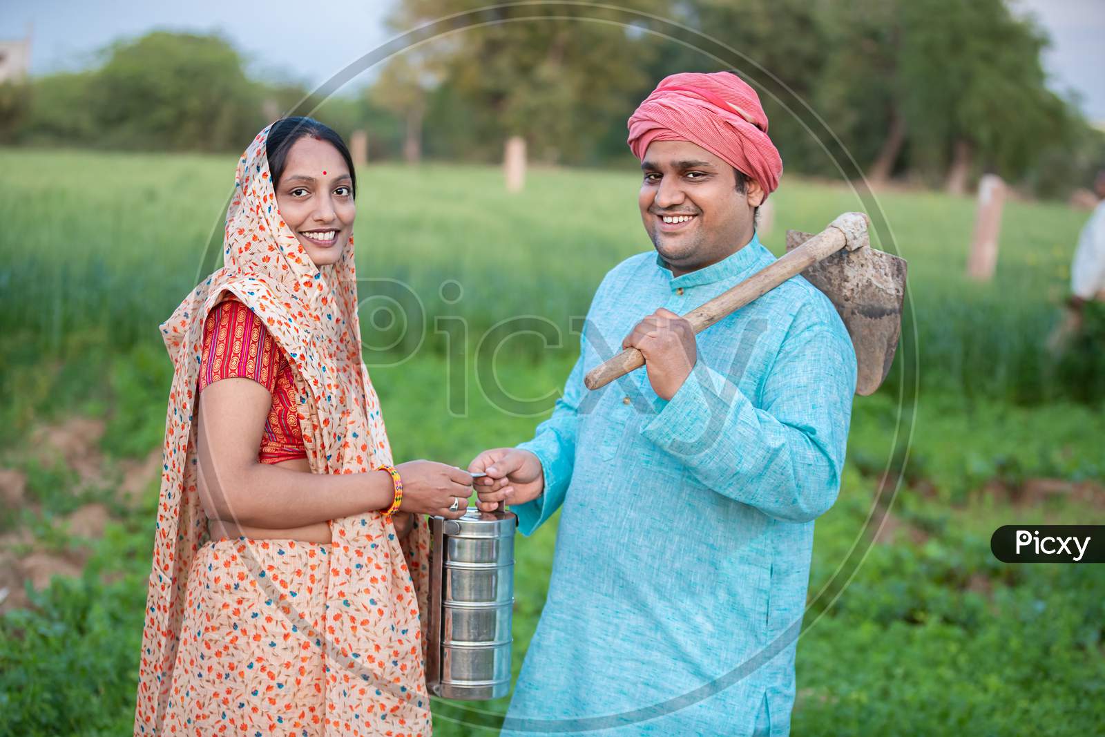 Portrait Of Traditional Indian Farmer Couple Or Labor Worker In Agriculture Field Holding Tiffing Lunch Box And Pretail Garden Spade / Shovel Tool.