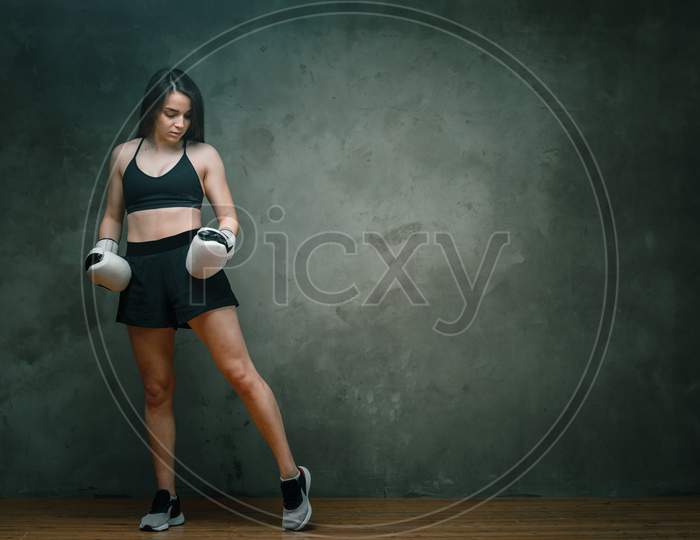 Young Athletic Woman Boxer In Shorts, Short Top And Boxing Gloves Posing  On Dark Gray Background. Full Length. Wall In Background.
