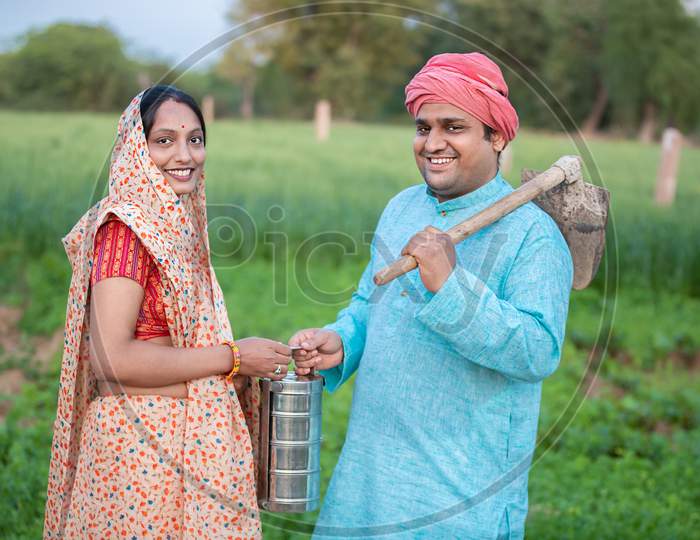 Portrait Of Traditional Indian Farmer Couple Or Labor Worker In Agriculture Field Holding Tiffing Lunch Box And Pretail Garden Spade / Shovel Tool.