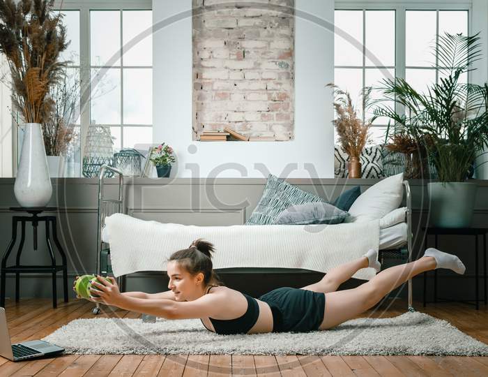 Positive Athletic Woman With Neatly Tied Hair And Tight Sportswear Holds The Plank  With Dumbbells On The Floor  At Home. The Young Woman Goes In For Sports At Home.