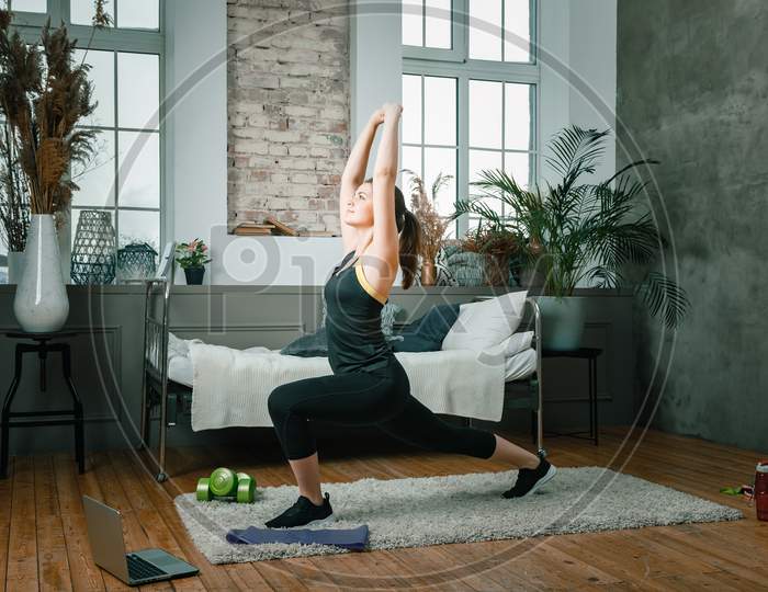 A Young Woman Goes In For Sports At Home, Online Workout From The Laptop. The Athlete Lunges, Watches A Movie  In The Bedroom, In The Background There Is A Bed, A Vase, A Carpet.