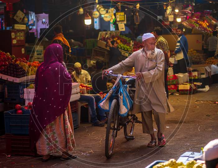 Muslim Old Age Man Wearing Traditional Islamic Cloths And Skull Cap Walking With Bicycle In A Fruit Market In South Mumbai