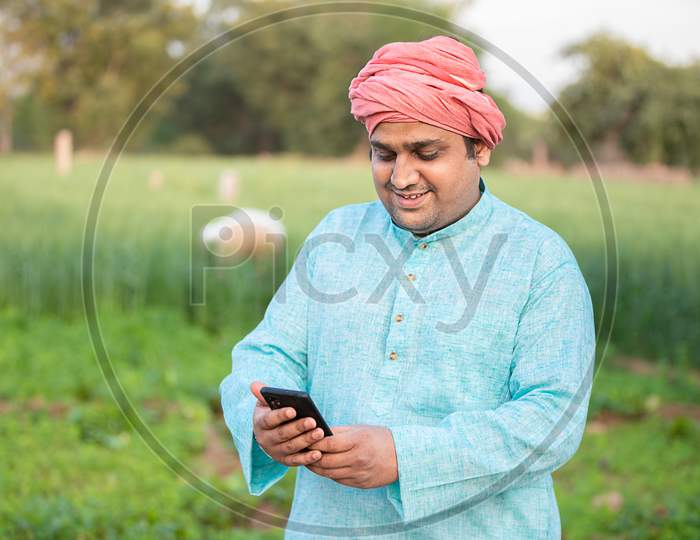 Young Happy Indian Farmer Worker Using Smartphone While Standing In Agriculture Green Field, Internet Banking, 5G Network Technology Concept, Male Wearing Traditional Kurta Outfit.