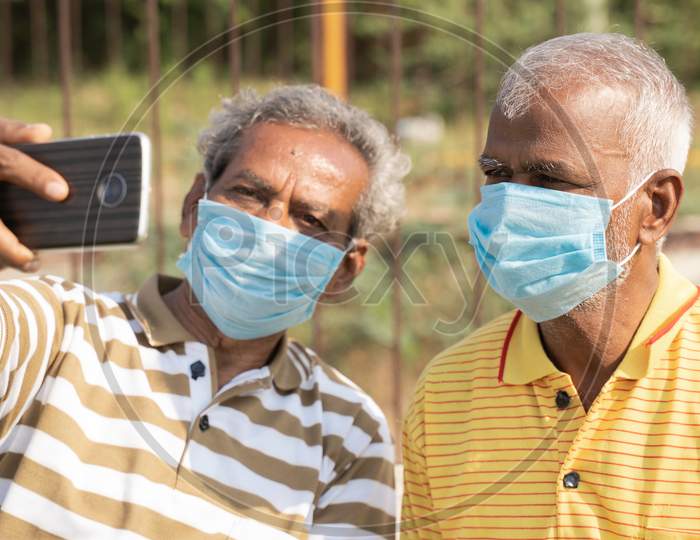 Elderly Friends With Medical Face Mask Taking Selfie While Sitting At Park - Concept Of Happy Senior People Friendship And Meetup After Lockdown Due To Coronavirus Covid-19 Pandemic.