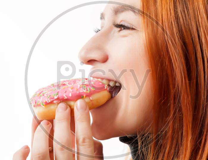 A Young Red-Haired Woman In A Black Turtleneck And A Kitchen Apron Laughs And Eats A Donut Pink With Pleasure On A White Isolated Background