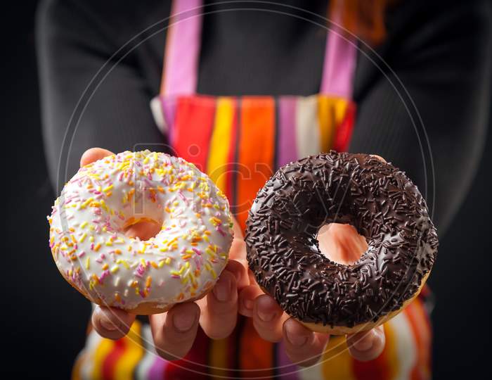 A Young Red-Haired Woman In A Black Turtleneck And Kitchen Apron Laughs And Shows What A Cool Chocolate Donuts Turned Out On A Black Isolated Background