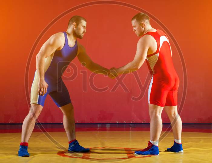 Two Greco-Roman  Wrestlers In Red And Blue Uniform Shake Hands Before Each Other  On A Yellow Wrestling Carpet In The Gym