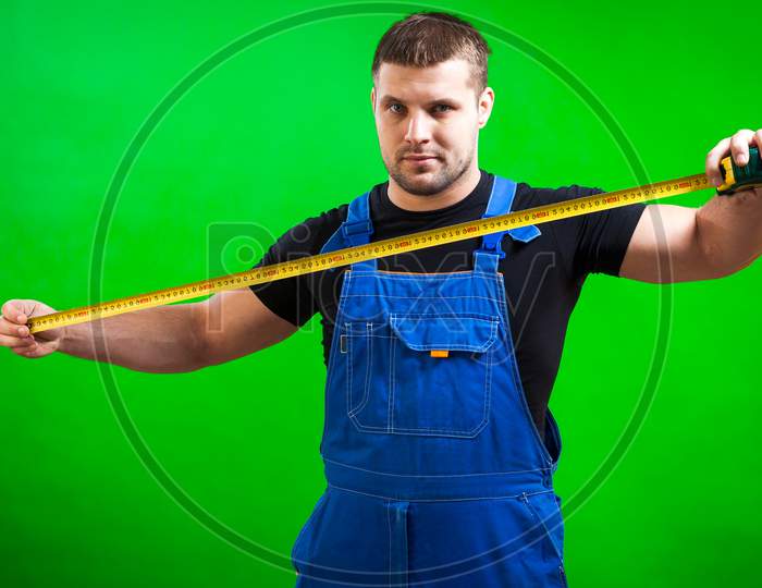 A Man Carpenter Wearing A Black T-Shirt And Blue Construction Jumpsuit Use A Long Yellow Tape Rule  On A Green Isolated Background