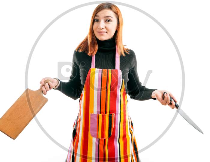 A Young Red-Haired Woman In A Black Turtleneck And Kitchen Apron Is Surprised And Holds A Large Kitchen Knife And A Wooden Kitchen Board For Cutting On A White Isolated Background