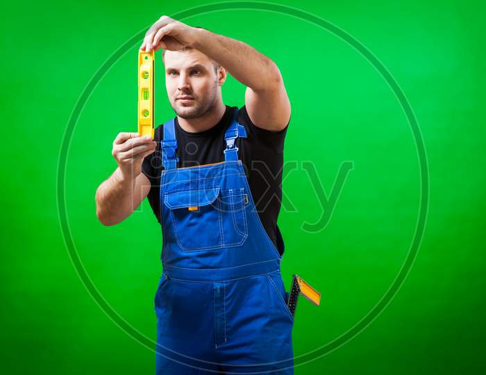 A Dark-Haired Male Construction Worker Wearing A Black T-Shirt And Blue Construction Overall Uses   Ruler Magnetic Level On A Green Isolated Background