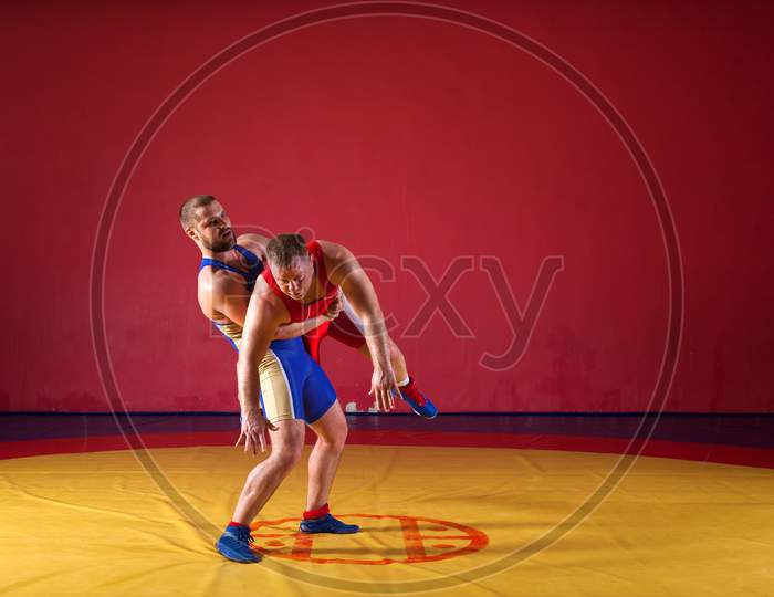 Two Strong Wrestlers In Blue And Red Wrestling Tights Are Wrestlng And Making A  Making A Hip Throw  On A Yellow Wrestling Carpet In The Gym. Young Man Doing Grapple.