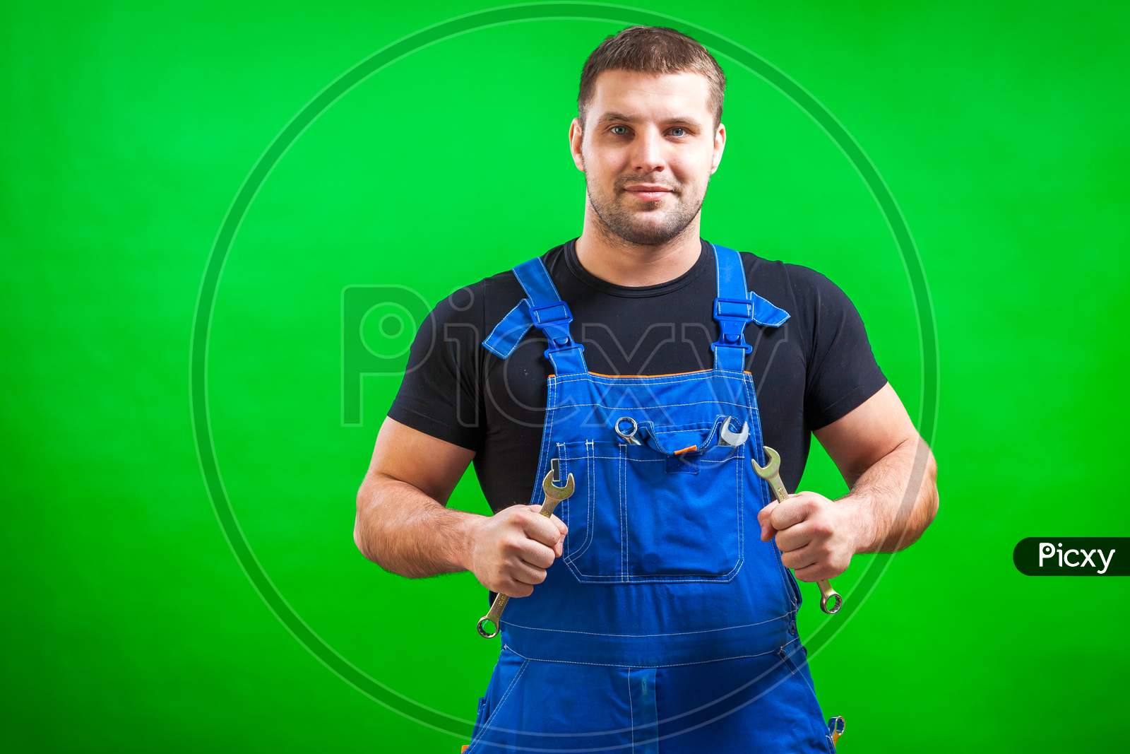 A Young Man Construction  Builder In A Black T-Shirt And Blue Construction Overall Holds In His Hands A New Tool Combination Wrench On A Green Isolated Background