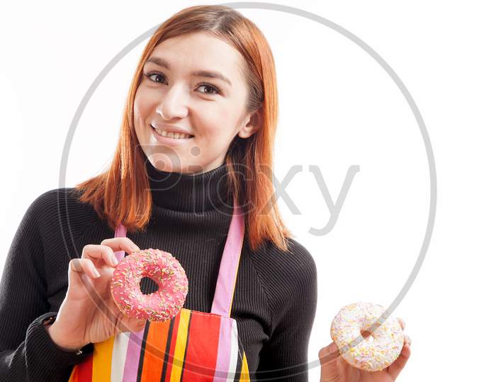 A Young Red-Haired Woman In A Black Turtleneck And A Kitchen Apron Laughs And Shows What A Cool Chocolate Donuts Are Made Of Pink And White On A White Isolated Background