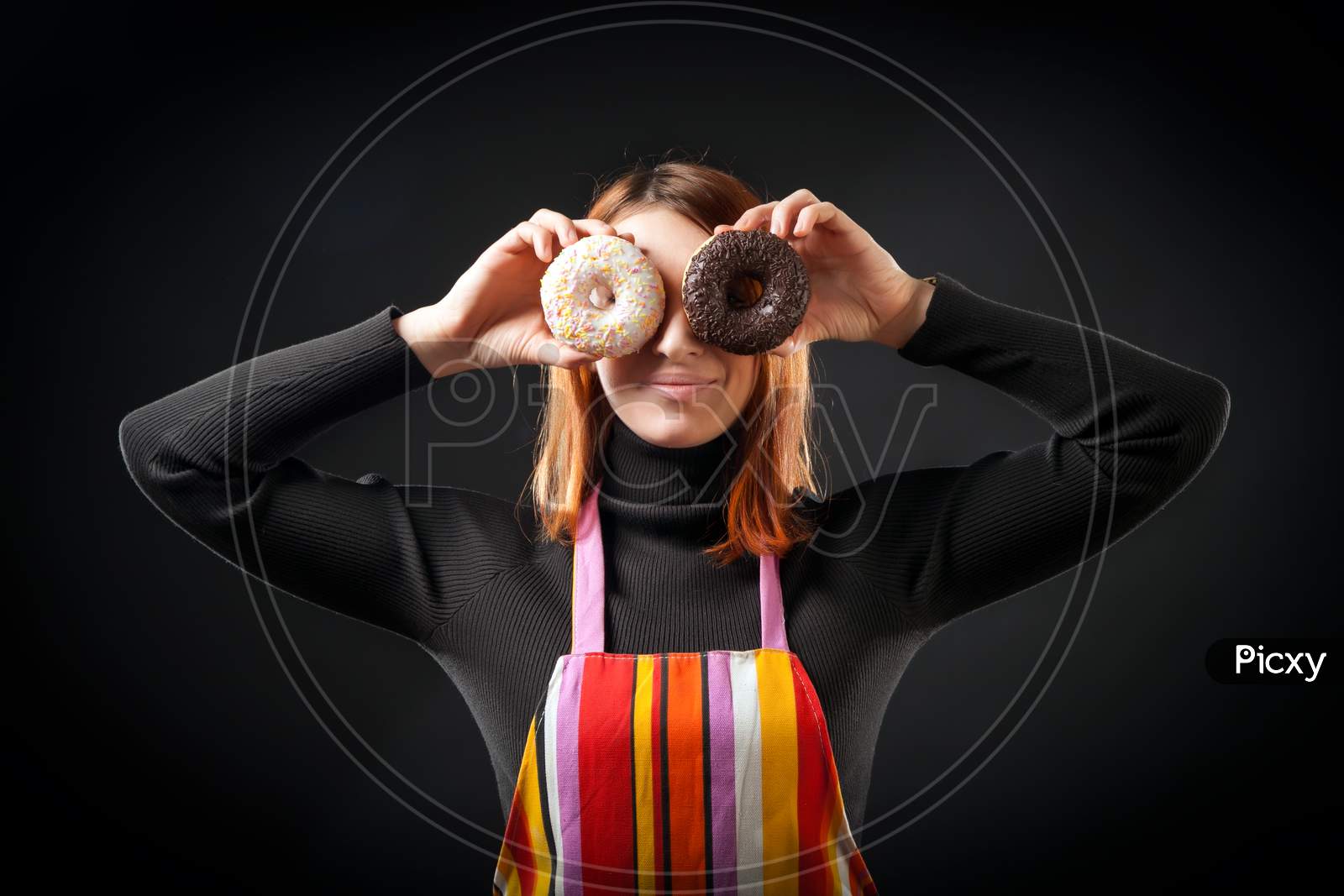 A Young Red-Haired Woman In A Black Turtleneck And Kitchen Apron Laughs And Holds Colorful Donuts As A Mask On A Black Isolated Background