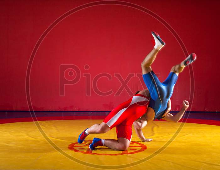 Two Young Men In Blue And Red Wrestling Tights Are Wrestlng And Making A Hip Throw On A Yellow Wrestling Carpet In The Gym