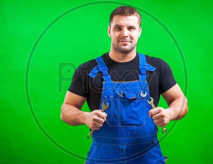 A Young Man Construction  Builder In A Black T-Shirt And Blue Construction Overall Holds In His Hands A New Tool Combination Wrench On A Green Isolated Background