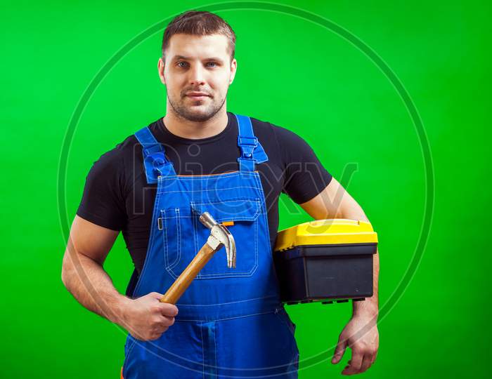 A Dark-Haired Male Construction Worker In A Black T-Shirt And Blue Construction Overall Holds A Wooden Hammer And A Box With Construction Tools On A Green Isolated Background