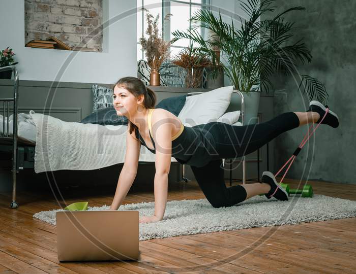 Positive Athletic Woman With Neatly Tied Hair And Tight Sportswear Doing Performs Lying Kicks With Sports Fitness Bands   At Home. The Young Woman Goes In For Sports At Home.