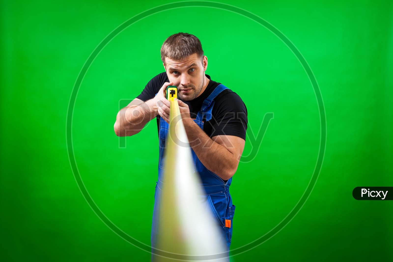 A Close-Up Of The Tape Rule. A Young Man Carpenter In A Black T-Shirt And Blue Construction Jumpsuit Measures The Length Of A Long Yellow Tape Rule For Construction Work On A Green Isolated Background