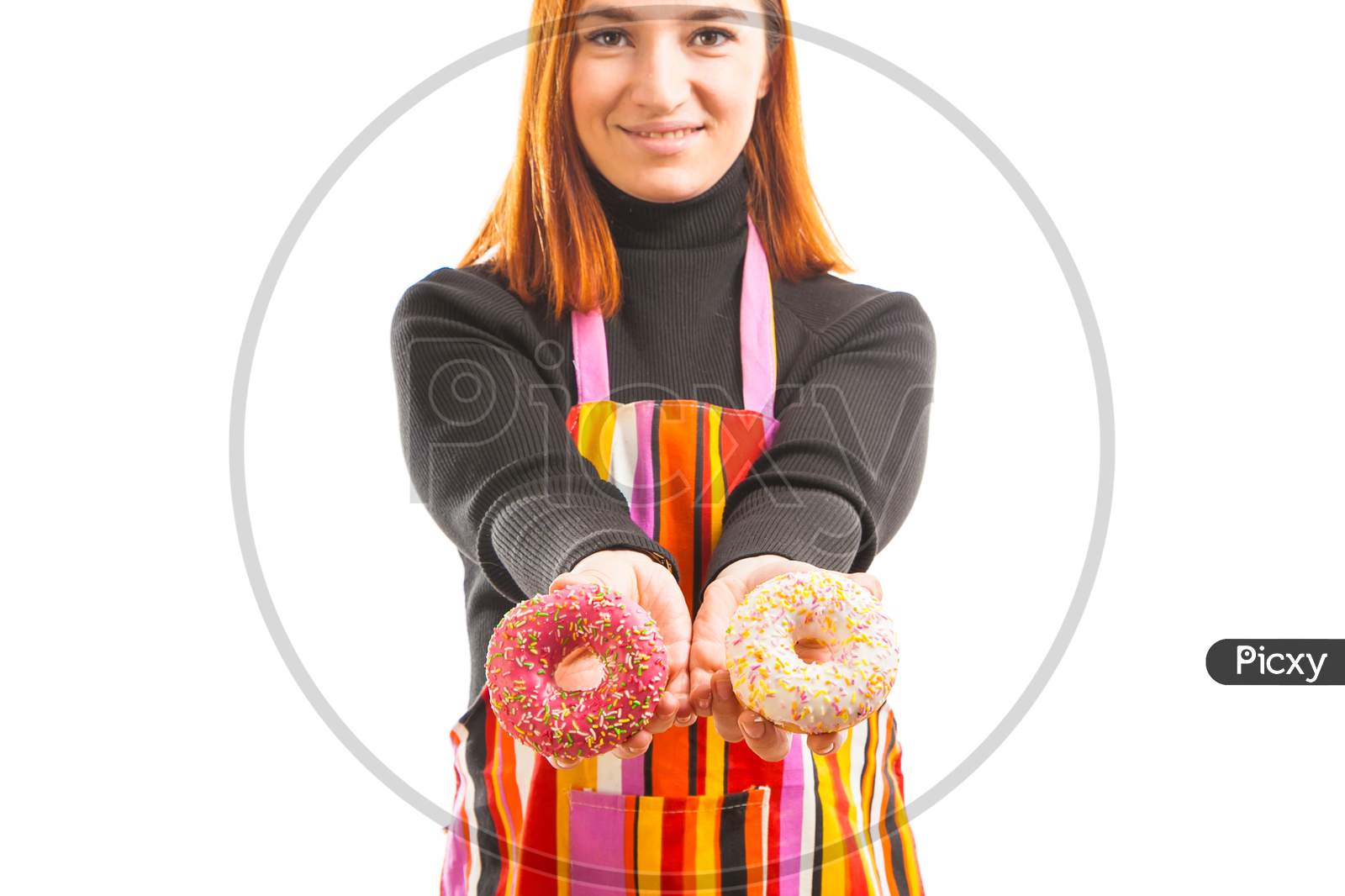 A Young Red-Haired Woman In A Black Turtleneck And Kitchen Apron Laughs And Shows What A Cool Chocolate Donuts Turned Out On A White Isolated Background
