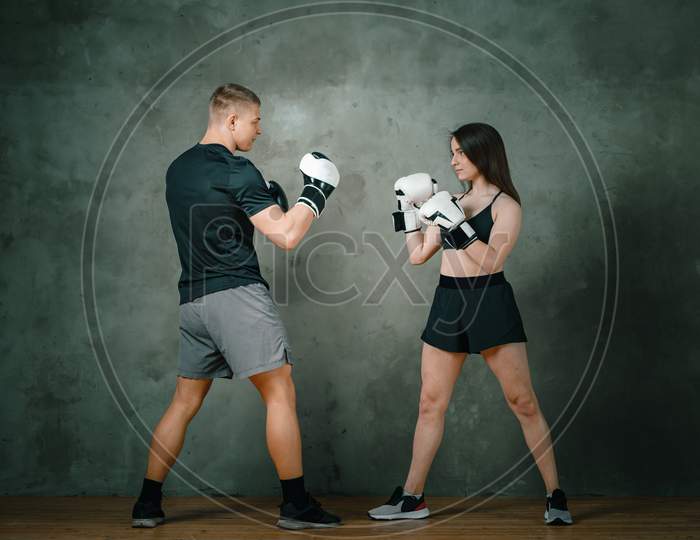Young Athletic Couple Of Man And Women Boxing In Boxing Gloves. A Woman Makes A Blow To The Head Of A Man On A Dark Gray Background. Full Length.
