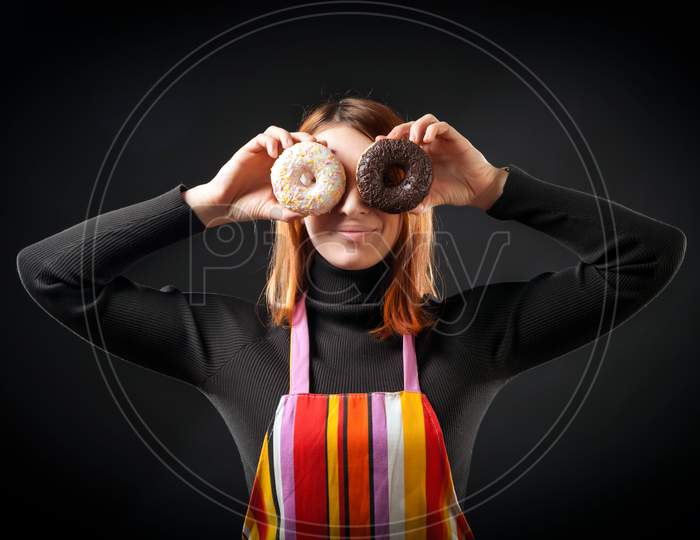 A Young Red-Haired Woman In A Black Turtleneck And Kitchen Apron Laughs And Holds Colorful Donuts As A Mask On A Black Isolated Background